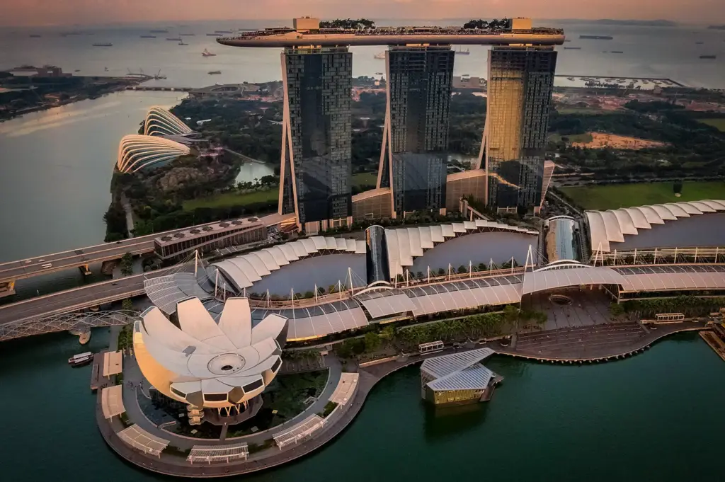 High-angle aerial view of Marina Bay Sands Singapore, showing the iconic three hotel towers, SkyPark, lotus-inspired ArtScience Museum, and Gardens by the Bay along Marina Bay at sunset