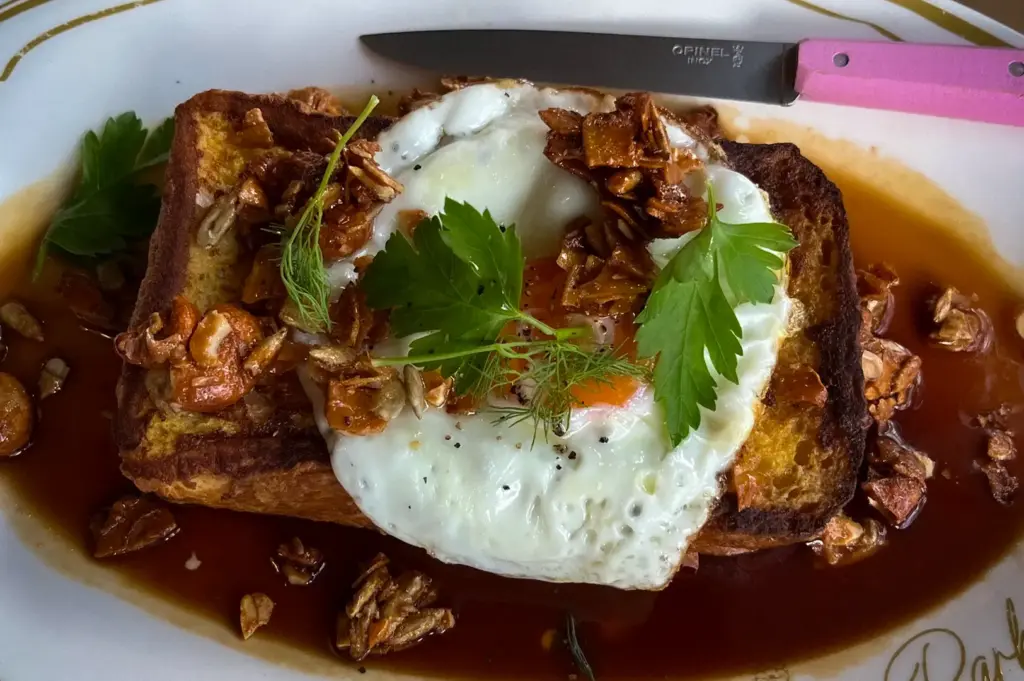 Savory French toast topped with a fried egg, crispy grains, and fresh herbs, served in a rich brown sauce on a branded Park Side Cafe plate with a pink knife