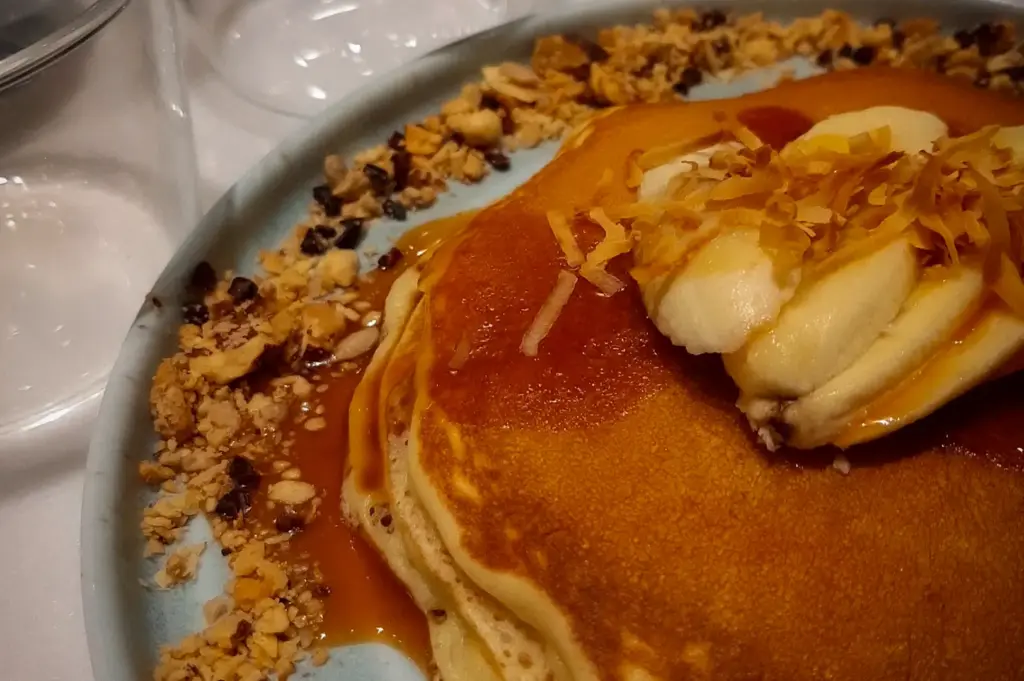 Stack of golden pancakes topped with sliced banana, toasted coconut flakes, syrup, and crushed nuts served on a ceramic plate at Acoustics Coffee Bar in Singapore